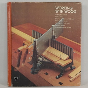 以下が含まれることがあります： 「Working with Wood」というタイトルの書籍で、ミトラスボックスとノコギリのクローズアップ画像が掲載されています。本の表紙は茶色で、白い文字が書かれています。ミトラスボックスは金属と木でできており、木片を保持しています。