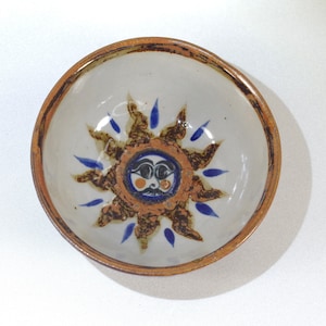 Puede incluir: Un tazón de cerámica blanca con un borde marrón y un diseño de sol pintado. El sol tiene una cara con ojos azules, una nariz negra y una boca azul. Los rayos del sol son marrones con puntas azules.