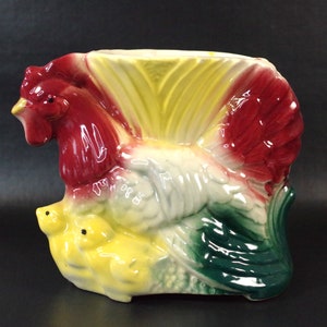 May include: A ceramic rooster planter with a red, white and green glaze. The rooster is holding two yellow chicks in its wings.
