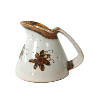 Puede incluir: Jarra de cerámica color crema con borde y asa marrones. La jarra presenta diseños florales marrones. Esta jarra es un artículo decorativo para la cocina o el comedor.