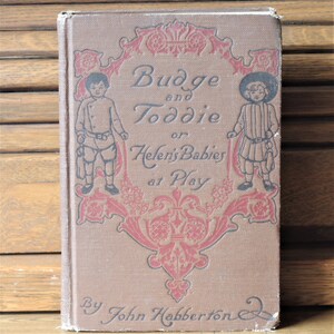 Pode incluir: Um livro vintage intitulado "Budge e Toddie ou os bebês de Helen a brincar" de John Habberton. A capa apresenta duas crianças com roupas antigas, com um design floral decorativo.