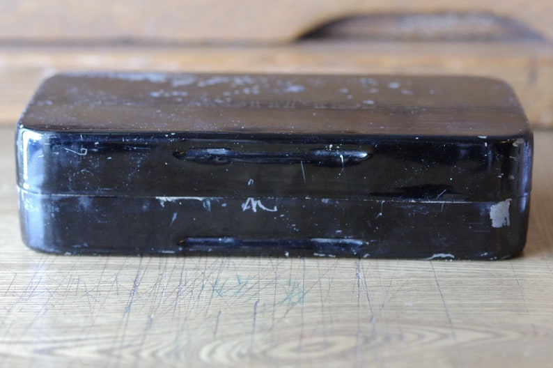 Pu&ograve; includere: Una scatola di metallo nera con un coperchio incernierato. La scatola ha due chiusure metalliche sul davanti e una finitura usurata e sbiadita.