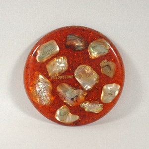 Może przedstawiać: Okrągły, pomarańczowy podkładka z wbudowanymi, opalizującymi kawałkami muszli. Podkładka ma napis "Yellowstone National Park" napisany złotymi literami.