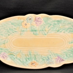 Op de afbeelding: Een vintage ovale serveerschaal van keramiek met een reliëfdesign van groene bladeren en pastelkleurige bloemen. De schaal heeft een crèmekleurige achtergrond.