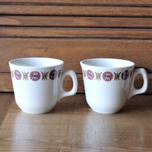 Può includere: Due tazze da caffè in ceramica bianca con una fascia decorativa di disegni floreali rosa, gialli e marroni. Ogni tazza ha un manico curvo e un bordo leggermente svasato. Le tazze sono appoggiate su una superficie di legno marrone chiaro.