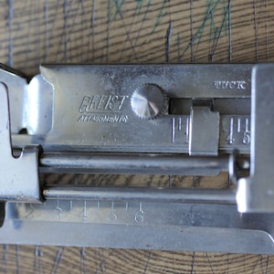 Pu&ograve; includere: Un accessorio per macchina da cucire argentato con un quadrante e un righello graduato in pollici. L'accessorio &egrave; etichettato "Greist Attachments" e "Tuck".