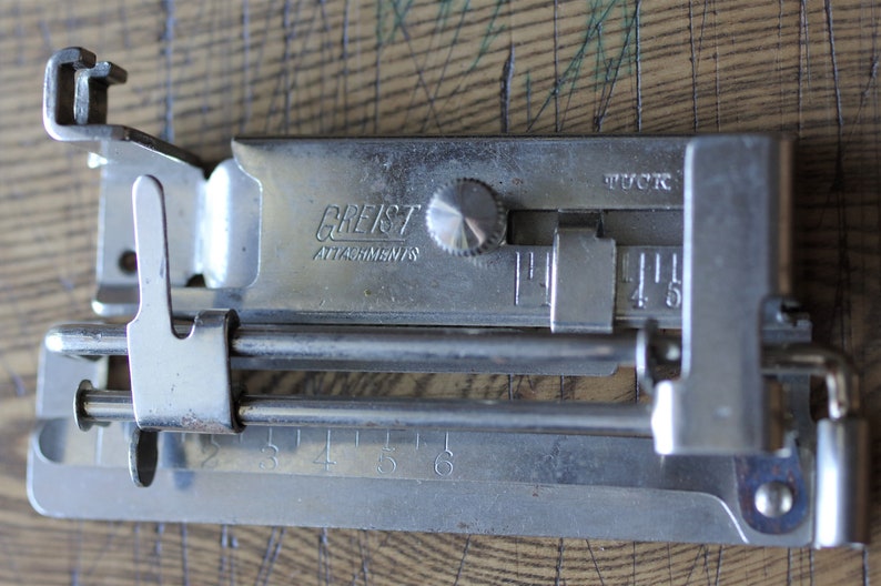 Pu&ograve; includere: Un accessorio per macchina da cucire argentato con un quadrante e un righello graduato in pollici. L'accessorio &egrave; etichettato "Greist Attachments" e "Tuck".