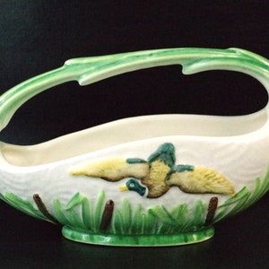 Puede incluir: Un macetero de cerámica blanca con forma de pato volando sobre juncos verdes. El macetero tiene un asa y está decorado con detalles verdes y marrones.