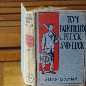 May include: A vintage book titled "Tom Fairfield's Pluck and Luck" by Allen Chapman. The cover features a boy in a suit holding a baseball bat and a suitcase. The book is part of the "Tom Fairfield Series" and published by Cupples & Leon Co.