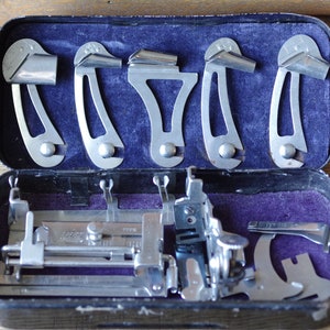 Pu&ograve; includere: Un kit di strumenti vintage per macchina da cucire in una custodia nera con fodera in velluto viola. Il kit include una variet&agrave; di strumenti in metallo, tra cui un piedino per macchina da cucire, un righello e un set di cinque guide in metallo.