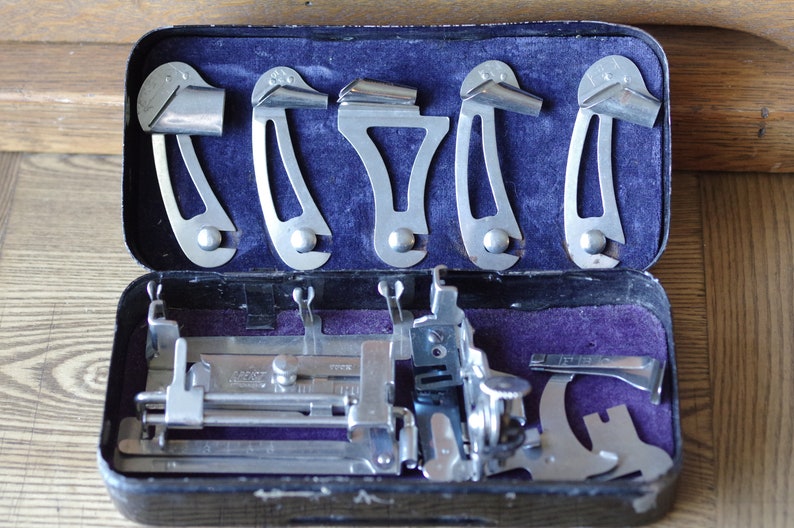 Pu&ograve; includere: Un kit di strumenti vintage per macchina da cucire in una custodia nera con fodera in velluto viola. Il kit include una variet&agrave; di strumenti in metallo, tra cui un piedino per macchina da cucire, un righello e un set di cinque guide in metallo.