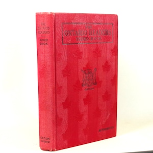 Op de afbeelding: Een rood hardback boek getiteld "The Ontario Readers Third Book" met een Canadees wapenschild en een esdoornbladontwerp. Het boek is uitgegeven door Eaton Toronto en is geautoriseerd.