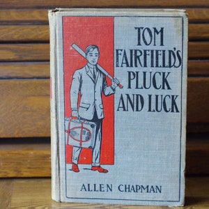 May include: A vintage book cover with a red and white background. The cover features a boy holding a baseball bat and a suitcase. The title of the book is "Tom Fairfield's Pluck and Luck" by Allen Chapman.