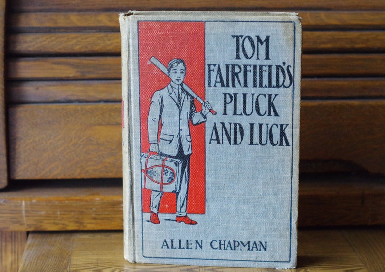 May include: A vintage book cover with a red and white background. The cover features a boy holding a baseball bat and a suitcase. The title of the book is "Tom Fairfield's Pluck and Luck" by Allen Chapman.