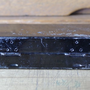 Pu&ograve; includere: Una scatola di metallo nera con una finitura usurata e sbiadita. La scatola ha due cerniere e un chiavistello sul davanti.