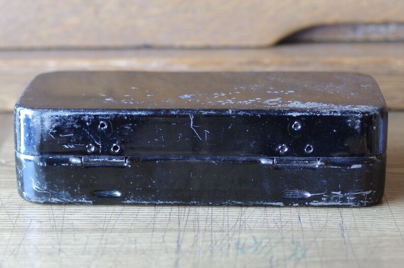 Pu&ograve; includere: Una scatola di metallo nera con una finitura usurata e sbiadita. La scatola ha due cerniere e un chiavistello sul davanti.