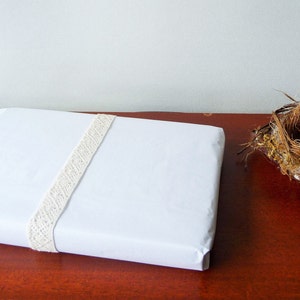 May include: A white gift box wrapped with a white lace ribbon. The box is sitting on a wooden surface.