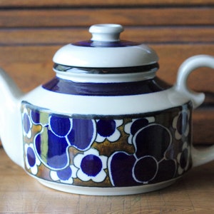 Pode incluir: Uma chaleira branca com um padrão floral azul e marrom. A chaleira tem uma tampa e uma alça.