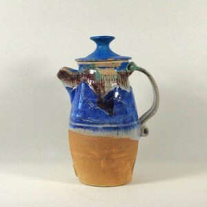 May include: A handmade ceramic teapot with a blue and brown glaze. The teapot has a lid and a handle. The teapot is sitting on a white surface.