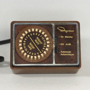 Puede incluir: Un temporizador de plástico marrón con un dial redondo que tiene los números del 1 al 12 y las palabras "DAY" y "NIGHT" en inglés y francés. El temporizador tiene un borde plateado y el texto "Engraham" en la parte delantera. El temporizador tiene tres ajustes: "On Marche", "Off Arrêt" y "Automatic".