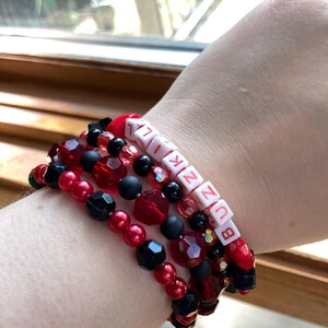May include: A stack of beaded bracelets in red, black, and clear beads. One bracelet features white letter beads spelling "BUNZIK". The bracelets are worn on a wrist.