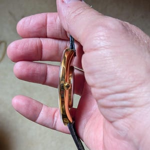 May include: A gold-toned wristwatch with a black leather strap. The watch face is round, and the case has a polished finish. The watch is held vertically, showcasing its side profile and the details of the band.