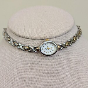 May include: A silver and gold-tone watch with a white face and gold hands. The watch is attached to a silver and gold-tone XOXO-style bracelet. The watch is displayed on a light pink mannequin head.