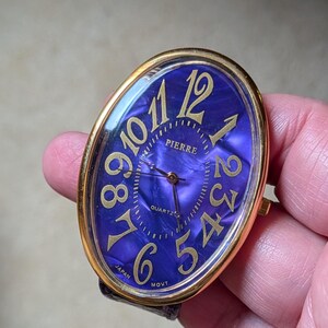 May include: Oval-shaped Pierre quartz watch with a gold-tone bezel and a deep purple face. The watch features gold numerals and hands, with the words "PIERRE", "QUARTZ", and "MOVT" visible. The watch band is black.