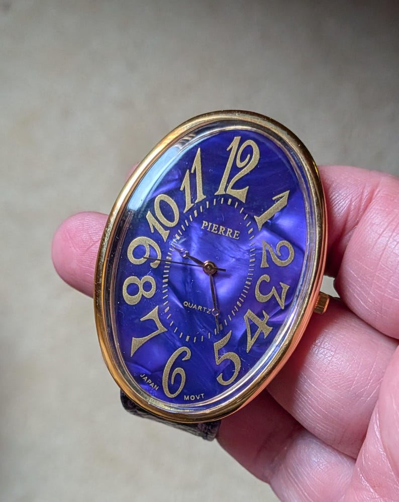 May include: Oval-shaped Pierre quartz watch with a gold-tone bezel and a deep purple face. The watch features gold numerals and hands, with the words "PIERRE", "QUARTZ", and "MOVT" visible. The watch band is black.