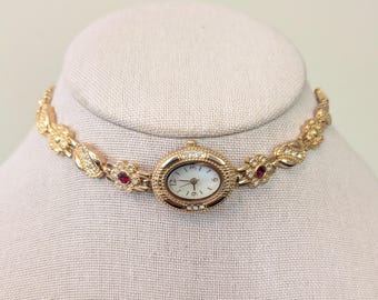 Collana girocollo vintage con orologio ovale senza marchio, color oro, cristalli rossi e dettagli floreali.