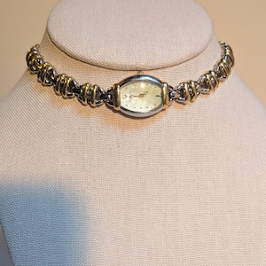 Puede incluir: Un collar de reloj plateado y dorado. La esfera del reloj es de forma ovalada con un fondo claro y manecillas e indicadores dorados. La correa está hecha de eslabones de metal plateado y dorado entrelazados.