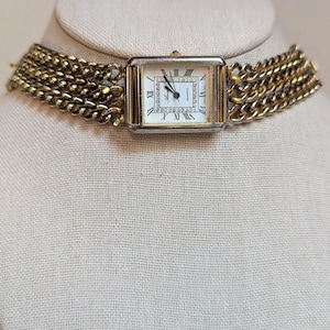 May include: A gold-tone watch with a square face and a chain link band. The watch face has Roman numeral hour markers and a white background. The watch is on a white mannequin.