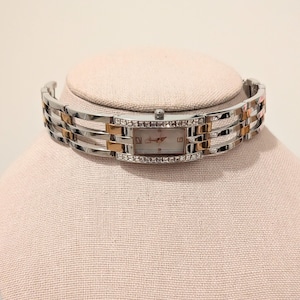May include: A silver and gold-toned rectangular wristwatch with a white face and crystal accents. The watch band is a combination of silver and gold links. The watch is displayed on a pink surface.