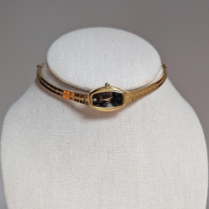 May include: A gold-coloured wristwatch with a black face and a small diamond accent. The watch is attached to a thin, gold-coloured band, and is displayed on a white mannequin head. The watch face is approximately 2 cm long.
