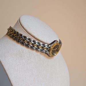 May include: A two-tone wristwatch with a silver and gold band and a black watch face. The watch is displayed on a neutral-colored mannequin head. The watch band is approximately 7 inches in length.