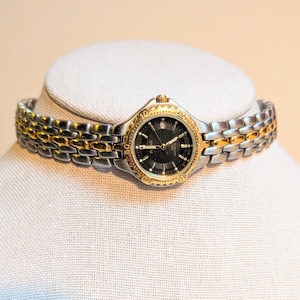 May include: A two-tone wristwatch with a black face and gold-tone bezel and accents. The watch has a silver and gold-tone link band. The watch is displayed on a white mannequin head.
