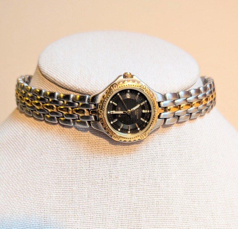 May include: A two-tone wristwatch with a black face and gold-tone bezel and accents. The watch has a silver and gold-tone link band. The watch is displayed on a white mannequin head.