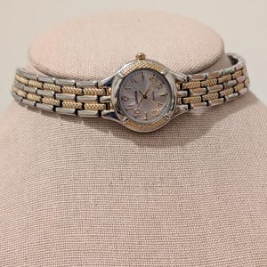 Puede incluir: Un reloj de cuarzo Geneva con una correa plateada y dorada. La esfera del reloj es de color claro con manecillas y números dorados. El reloj se exhibe en una cabeza de maniquí de color neutro.