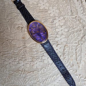 May include: An oval-faced wristwatch with a gold-tone bezel and a dark purple face with gold numerals. The watch has a black snakeskin-patterned band with a gold buckle. The watch is displayed on a cream-colored, textured surface.