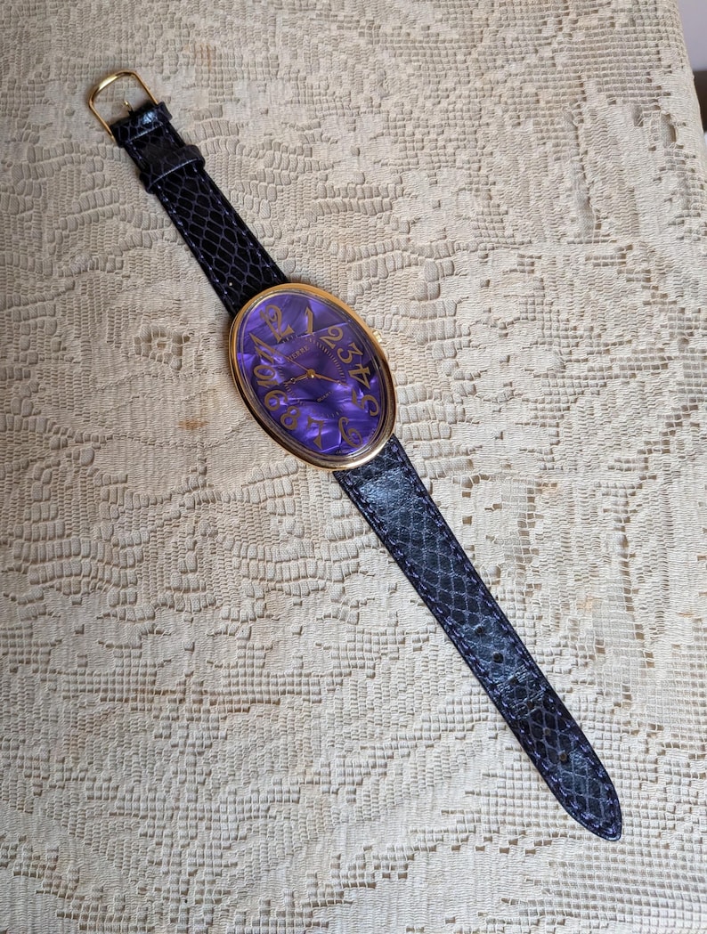 May include: An oval-faced wristwatch with a gold-tone bezel and a dark purple face with gold numerals. The watch has a black snakeskin-patterned band with a gold buckle. The watch is displayed on a cream-colored, textured surface.