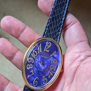May include: A gold-toned Pierre watch with a blue face and gold numerals. The watch has a black, textured band and a gold buckle. The watch face is oval-shaped and features the word "Pierre".