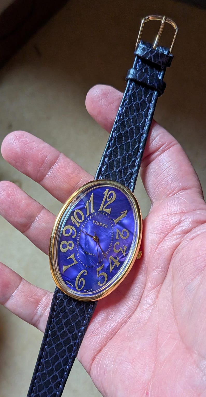 May include: A gold-toned Pierre watch with a blue face and gold numerals. The watch has a black, textured band and a gold buckle. The watch face is oval-shaped and features the word "Pierre".