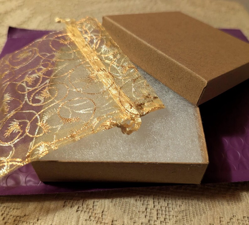 May include: An open brown cardboard box with a matching lid, revealing a white interior. A gold mesh bag with an ornate design is partially inside the box. The box sits on a purple padded envelope.