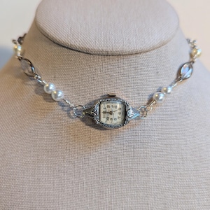 Puede incluir: Un collar de estilo vintage con un reloj plateado como pieza central. El reloj tiene una esfera color crema con números negros. El collar está adornado con perlas blancas y cuentas ovaladas transparentes, creando un accesorio elegante y único.