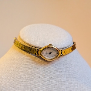 May include: A gold-toned wristwatch with an oval face and a matching gold band. The watch face is white with black numerals and hands. The band is approximately 0.5 inches wide.