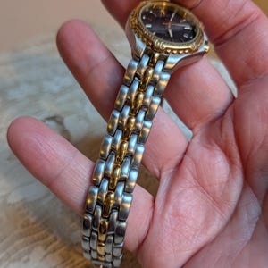 May include: A two-tone wristwatch with a silver and gold band. The watch face is dark with gold accents and hands. The watch is held in a hand, showcasing the details of the band and face.