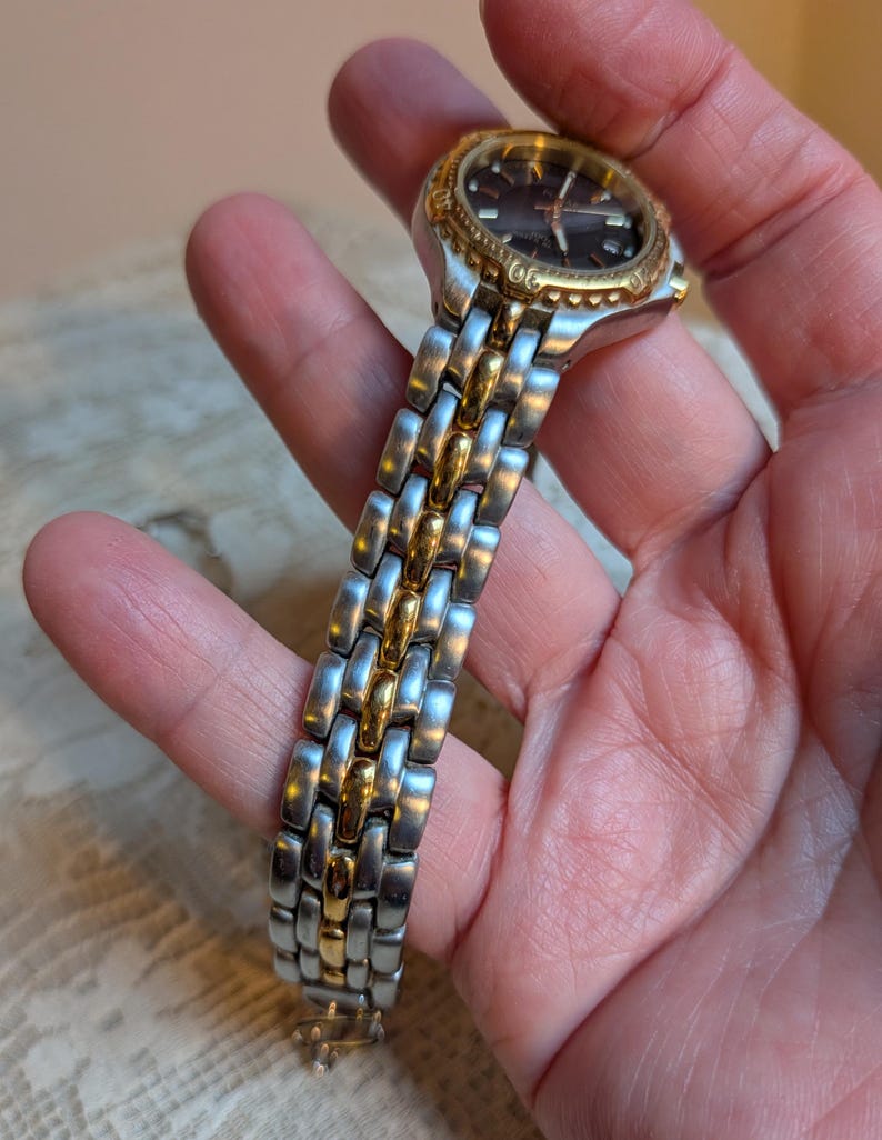 May include: A two-tone wristwatch with a silver and gold band. The watch face is dark with gold accents and hands. The watch is held in a hand, showcasing the details of the band and face.