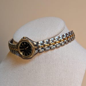 May include: A two-tone wristwatch with a silver and gold band and a black watch face. The watch is displayed on a neutral-colored mannequin head. The watch band is approximately 0.5 inches wide.