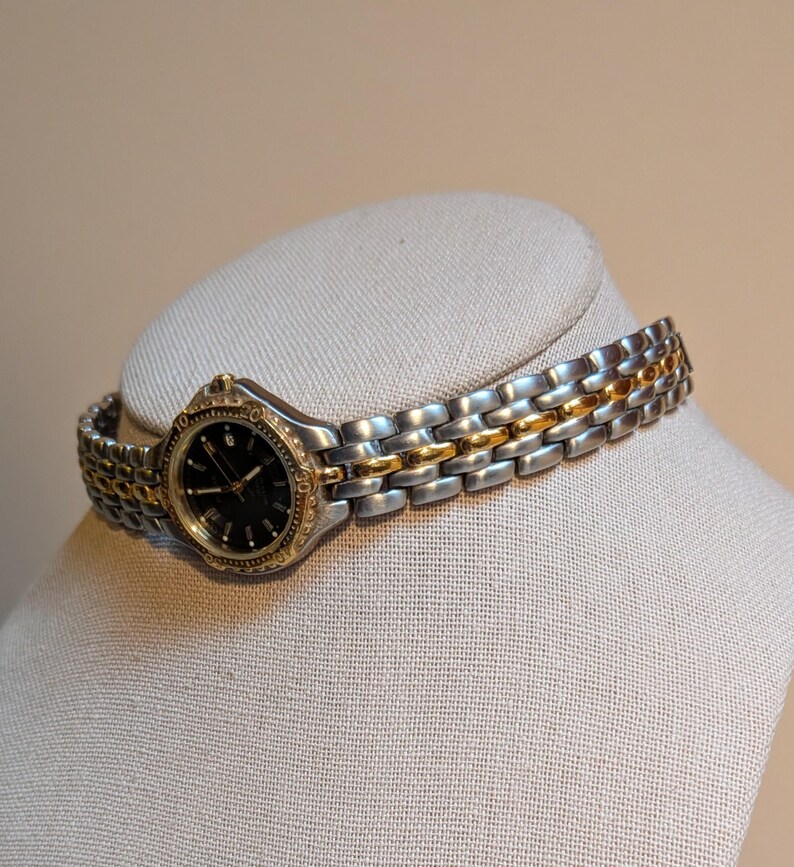 May include: A two-tone wristwatch with a silver and gold band and a black watch face. The watch is displayed on a neutral-colored mannequin head. The watch band is approximately 0.5 inches wide.