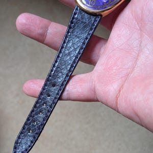 May include: A gold-toned wristwatch with a purple face and black leather band. The watch face has gold numerals and a swirling pattern. The black leather band has a textured, snake-like pattern.